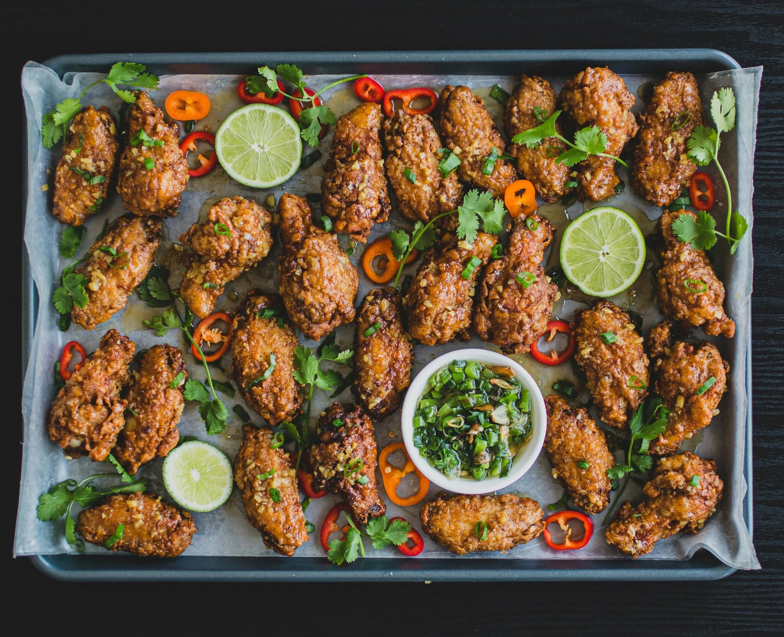 Baked chicken wings with lime slices, red peppers, and a small bowl of green sauce on a baking tray.