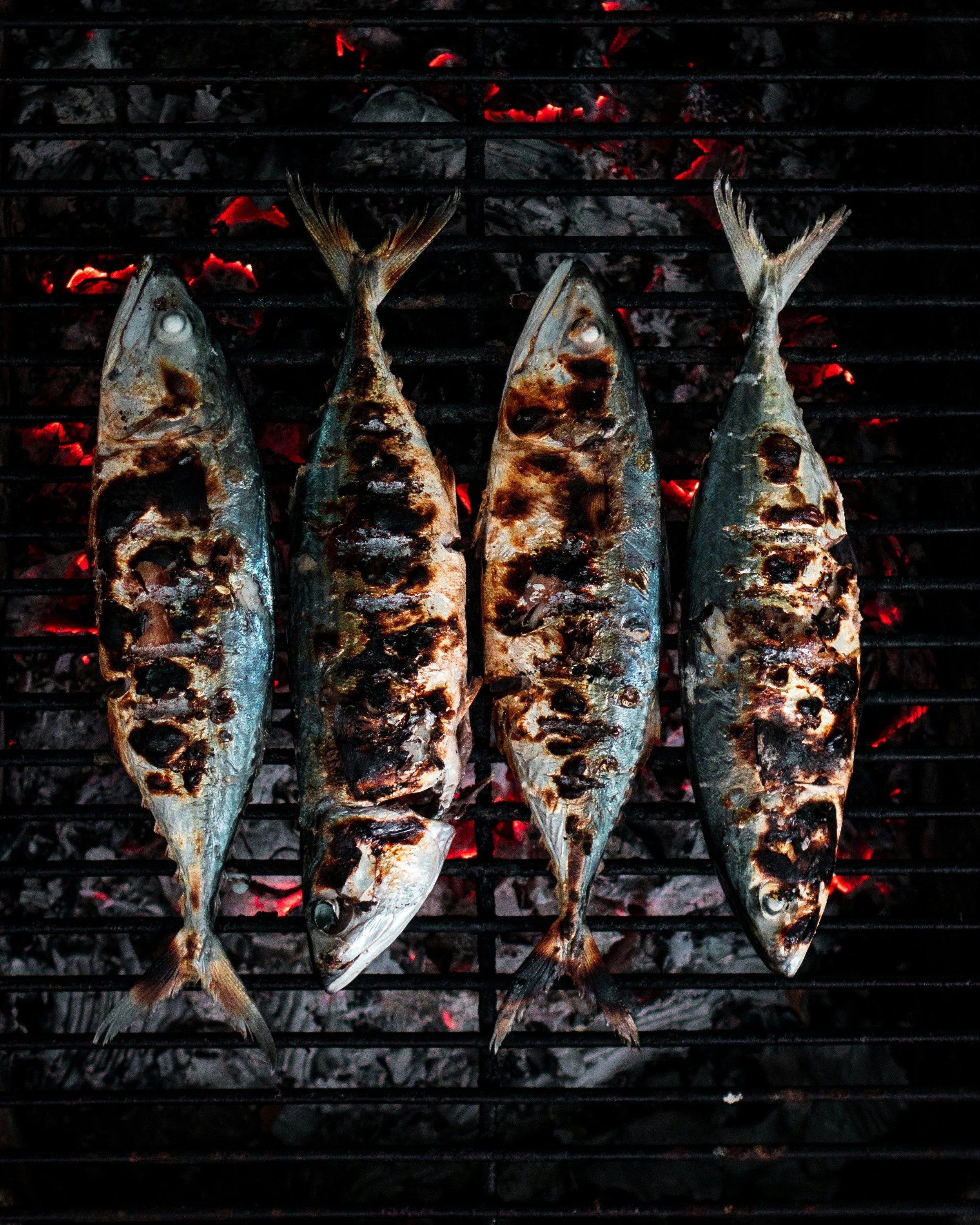 Four fish being grilled on a barbecue with visible flames and coals.
