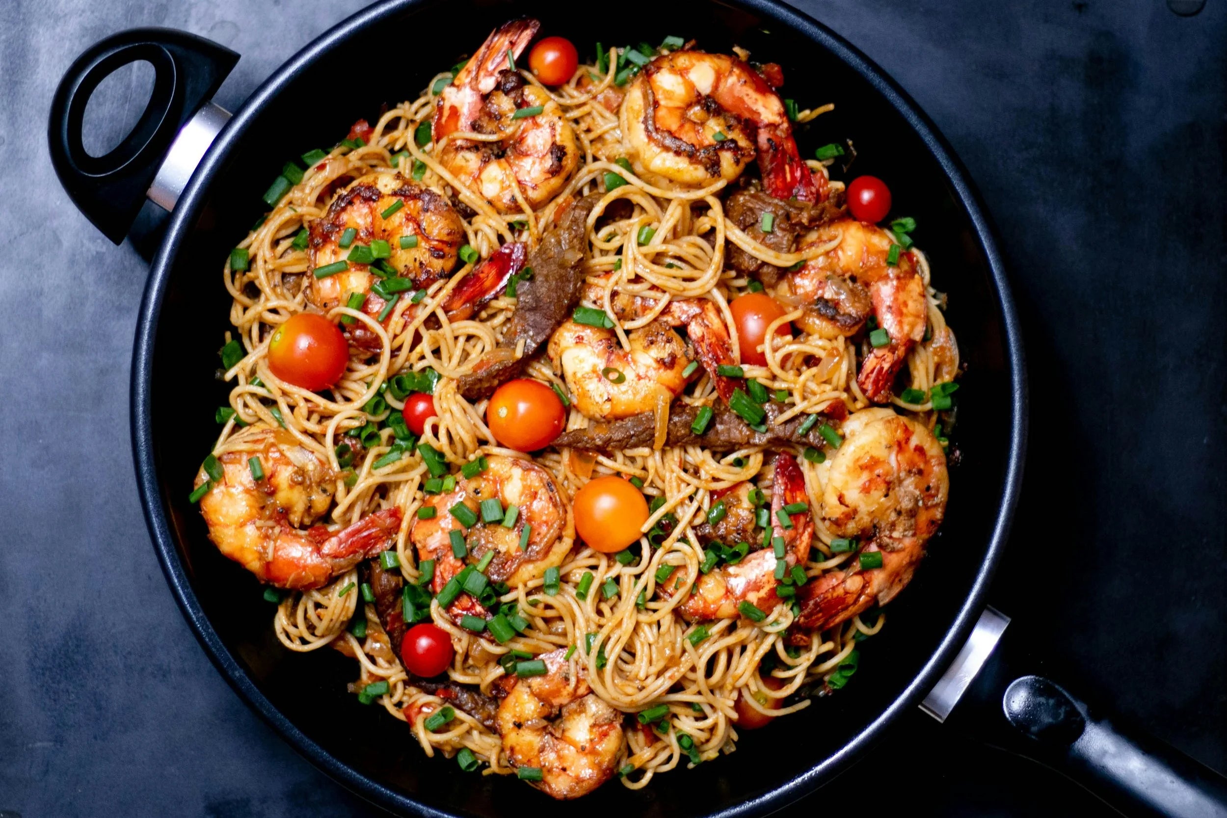Fried noodles with shrimp and vegetables in a black pan on a dark background