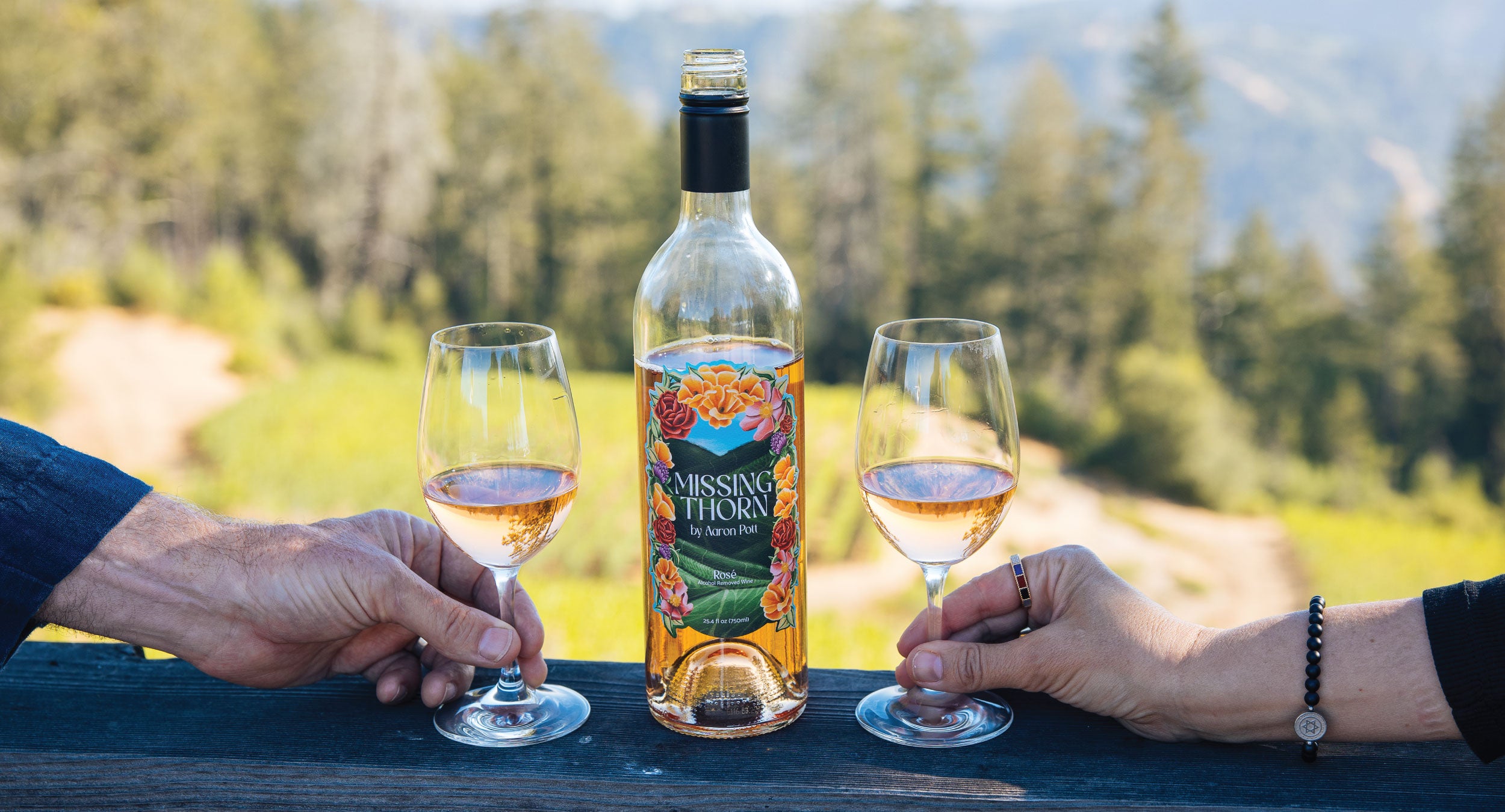 Two people holding wine glasses with a bottle of 'Missing Thorn' wine against a scenic background.