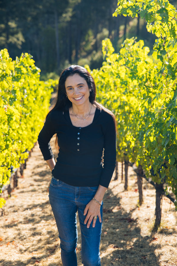Stephanie Honig smiling in sunny vineyard  framed by rows of green grapevines.